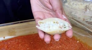 Hand holding stuffed pasta shell with creamy filling over tomato sauce in a baking dish.